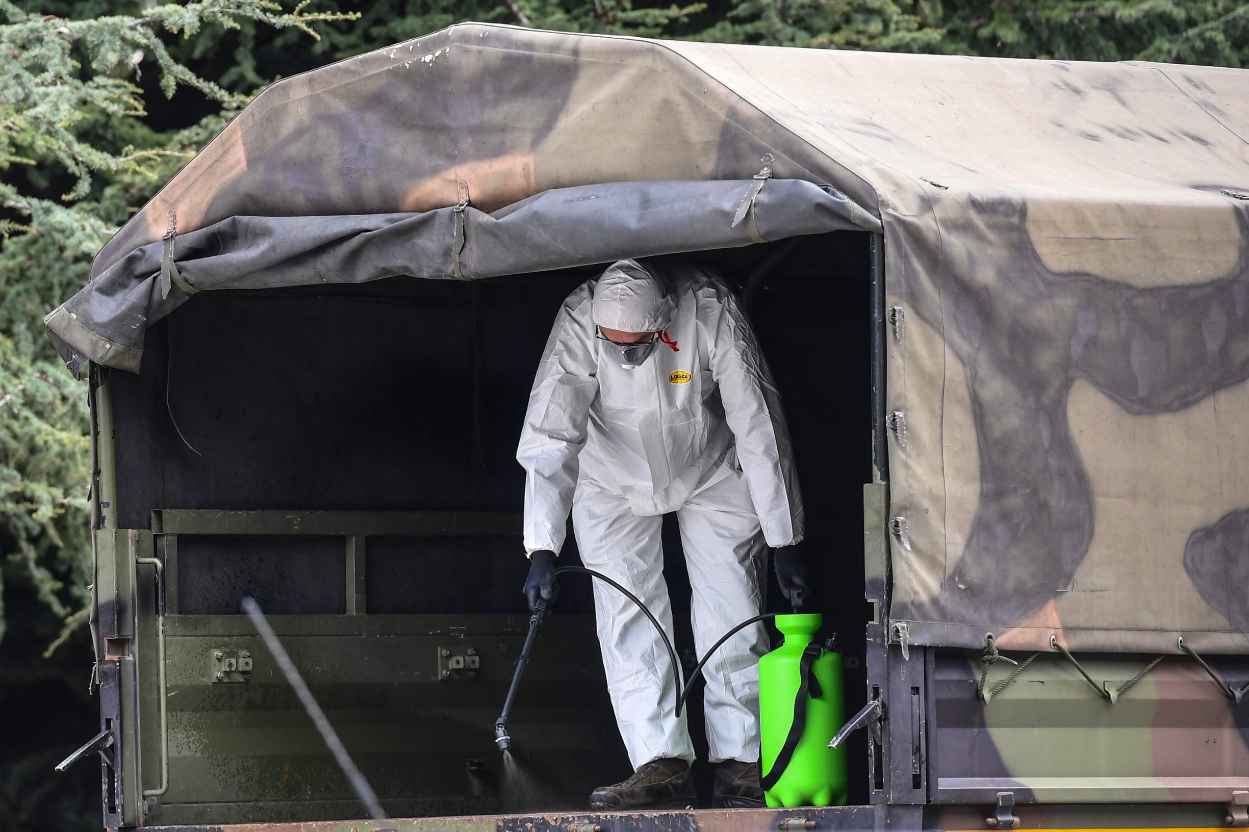 CORRECTION - A man wearing a protective gear sprays disinfectant in one of the Italian Army trucks parked on March 26, 2020 at the Monumental Cemetery of Bergamo, Lombardy, waiting to load coffins of victims of the coronavirus, to bring them to crematoriums in other regions where municipalities have made themselves available to accept them, during the country's lockdown following the COVID-19 new coronavirus pandemic. (Photo by Miguel MEDINA / AFP) / The erroneous mention appearing in the metadata of this photo by Miguel MEDINA has been modified in AFP systems in the following manner: [Wait to laod coffins] instead of [bringing back urns containing ashes]. Please immediately remove the erroneous mention from all your online services and delete it from your servers. If you have been authorized by AFP to distribute it to third parties, please ensure that the same actions are carried out by them. Failure to promptly comply with these instructions will entail liability on your part for any continued or post notification usage. Therefore we thank you very much for all your attention and prompt action. We are sorry for the inconvenience this notification may cause and remain at your disposal for any further information you may require.