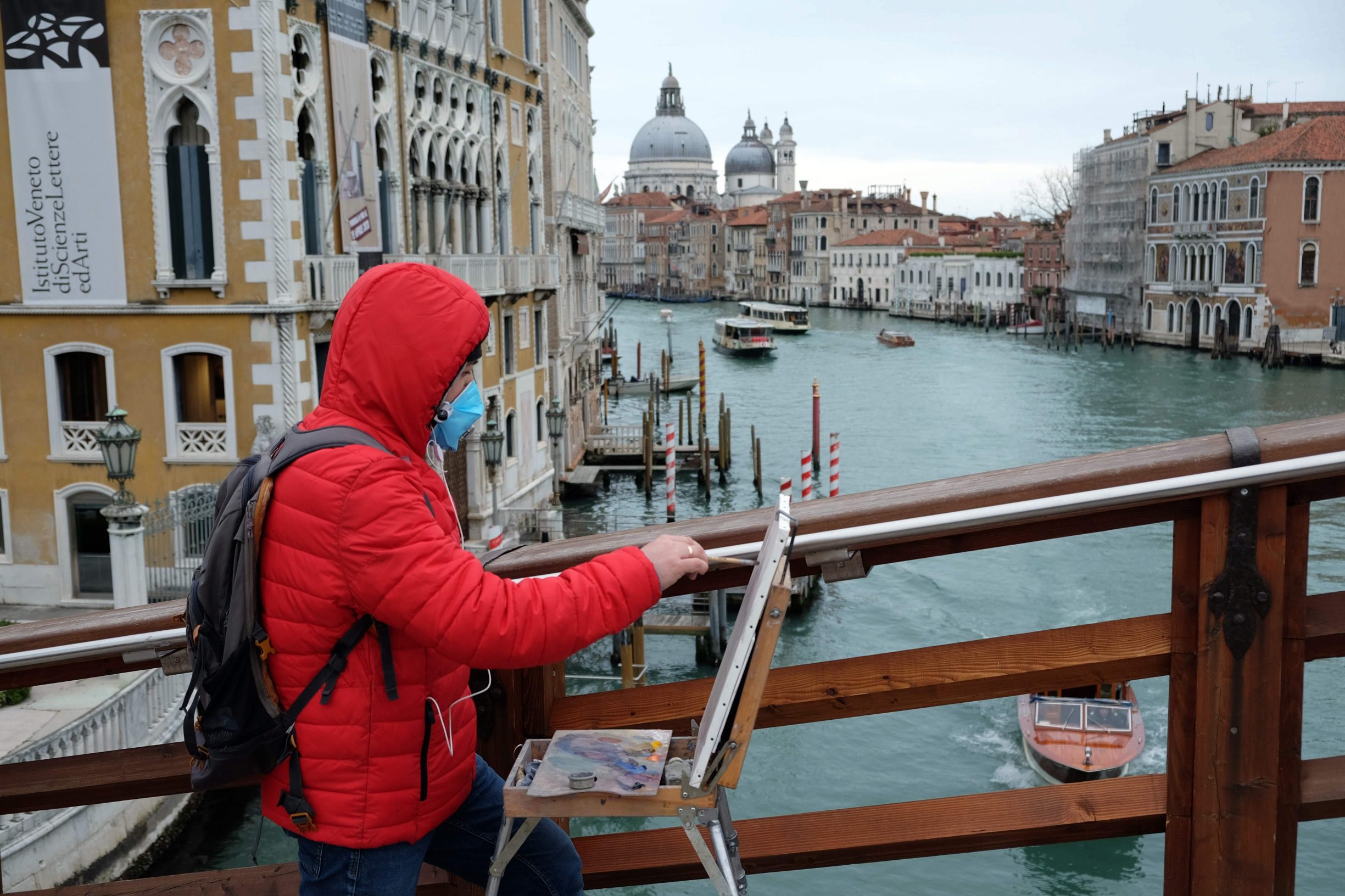 A man wearing a protective mask paint on Accademia bridge in Venice on March 8, 2020 on March 8, 2020 in Venice shows the Grand Canal empty of boats with the Salute Church on the left. - A quarter of the Italian population was locked down on March 8, 2020 as the government takes drastic steps to stop the spread of the deadly new coronavirus that is sweeping the globe, with Latin America recording its first fatality. (Photo by ANDREA PATTARO / AFP)