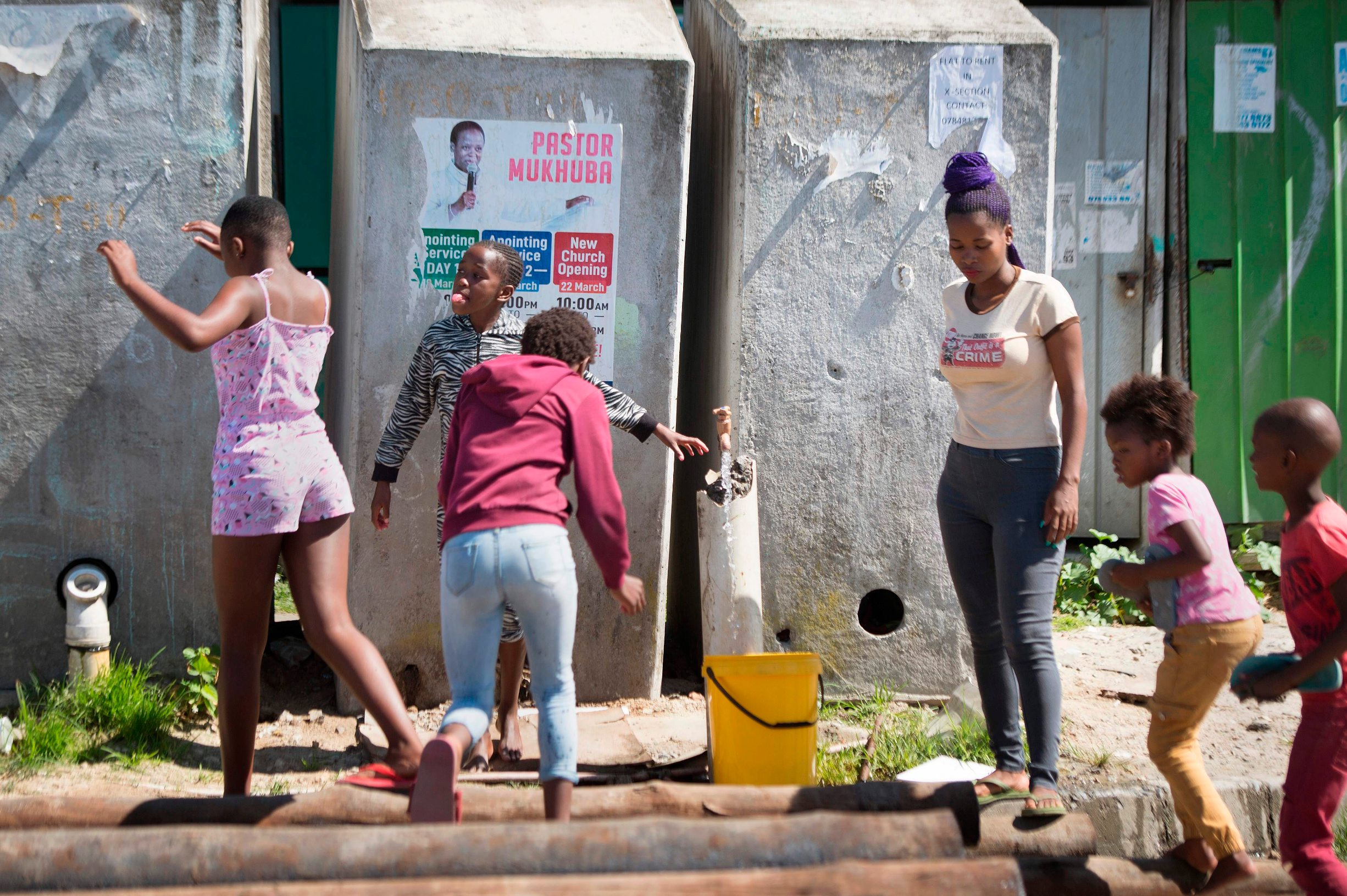 A woman fills a bucket from a communal tap as children play  in front of a line of communal toilets in Khayelitsha, near Cape Town, on March 31, 2020 in Cape Town. - Local government administrators in Cape Town said on March 29, 2020, a COVID-19 coronavirus case had been detected in Khayelitsha, the city's largest township, where hundreds of thousands live in shacks. 
An outbreak in the crowded townships where water and sanitation are problematic, could prove difficult to contain in the country which already has the highest number of infections in Africa. (Photo by RODGER BOSCH / AFP)