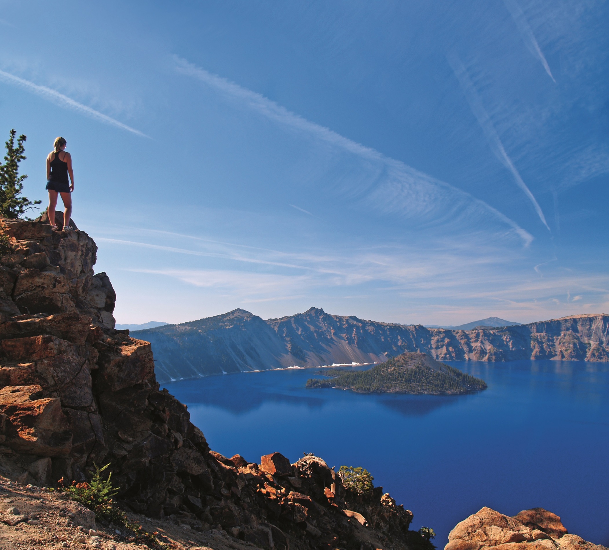 Girl looks out over the expanse of the deep blues of Crater Lake standing on a precarious rock outcrop