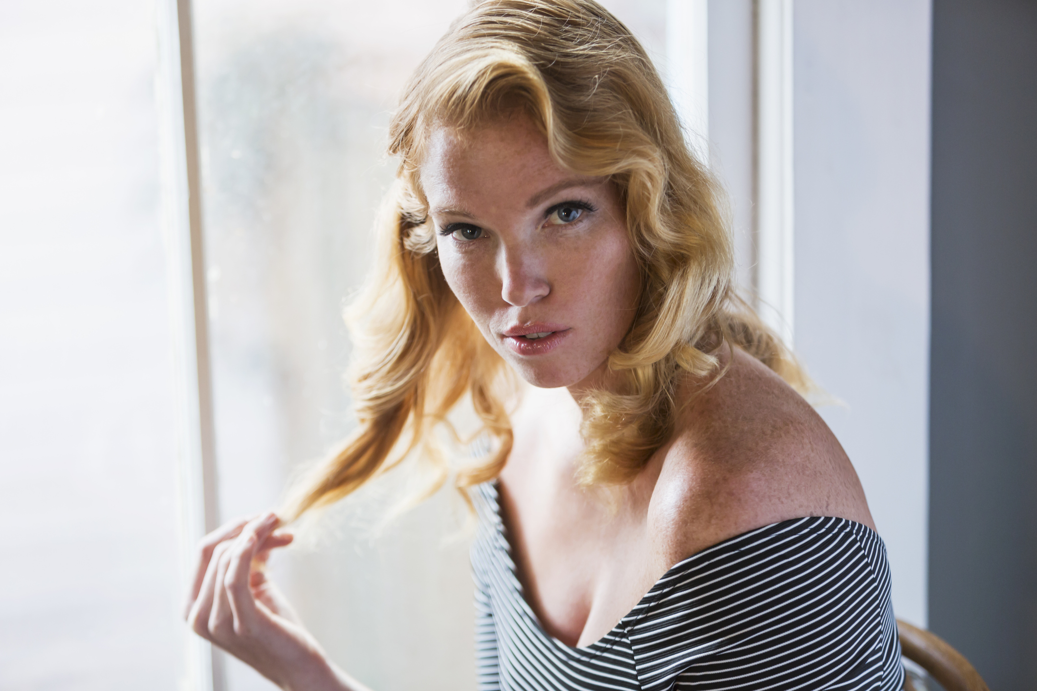 Seductive young blonde woman sitting indoors by sunny window, staring at the camera, playing with her hair.