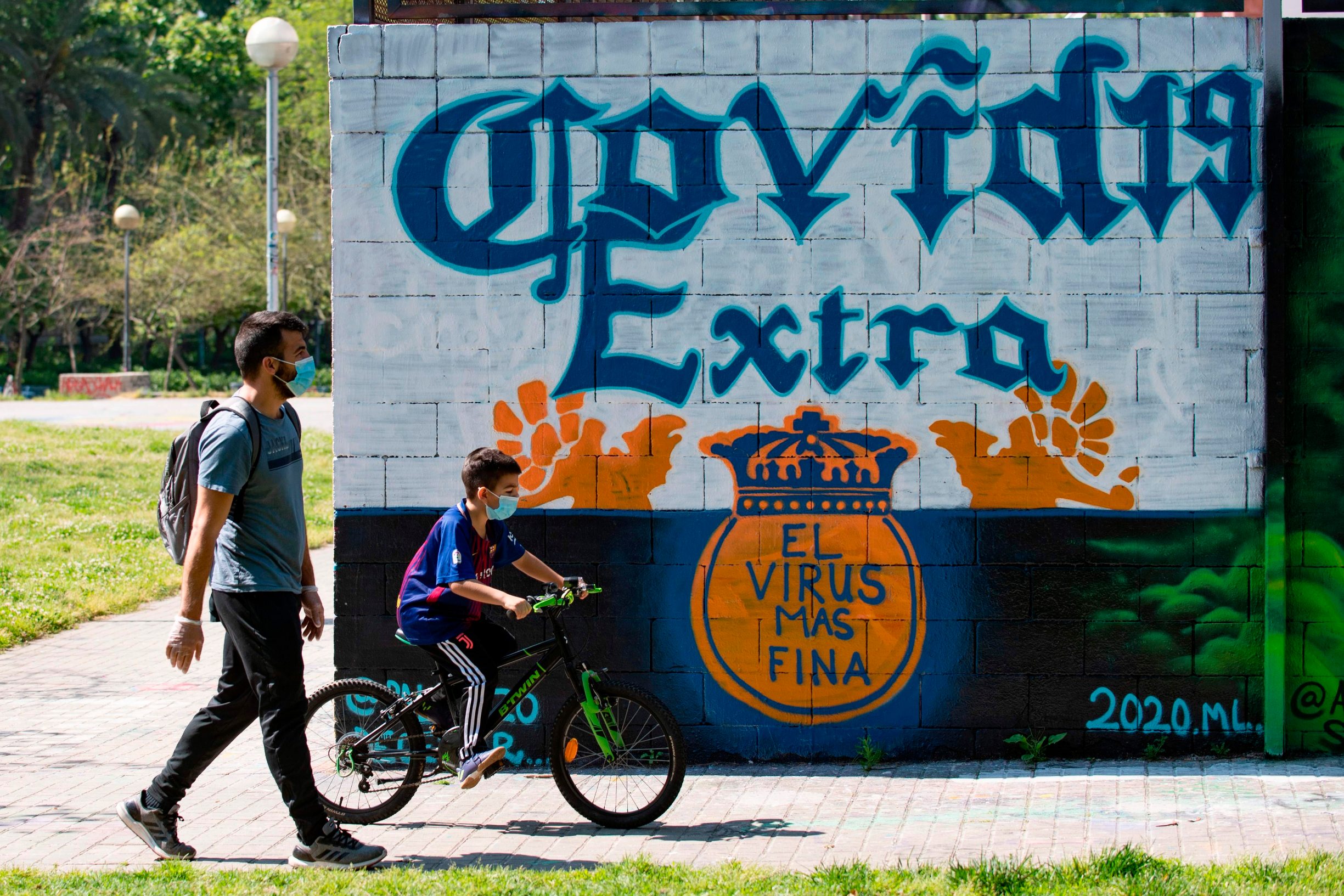 A boys rides his bike beside his father as they walk past a COVID-19 graffiti in Barcelona  on April 26, 2020 amid a national lockdown to prevent the spread of the COVID-19 disease. - After six weeks stuck at home, Spain's children were being allowed out today to run, play or go for a walk as the government eased one of the world's toughest coronavirus lockdowns. Spain is one of the hardest hit countries, with a death toll running a more than 23,000 to put it behind only the United States and Italy despite stringent restrictions imposed from March 14, including keeping all children indoors. Today, with their scooters, tricycles or in prams, the children accompanied by their parents came out onto largely deserted streets. (Photo by Josep LAGO / AFP)