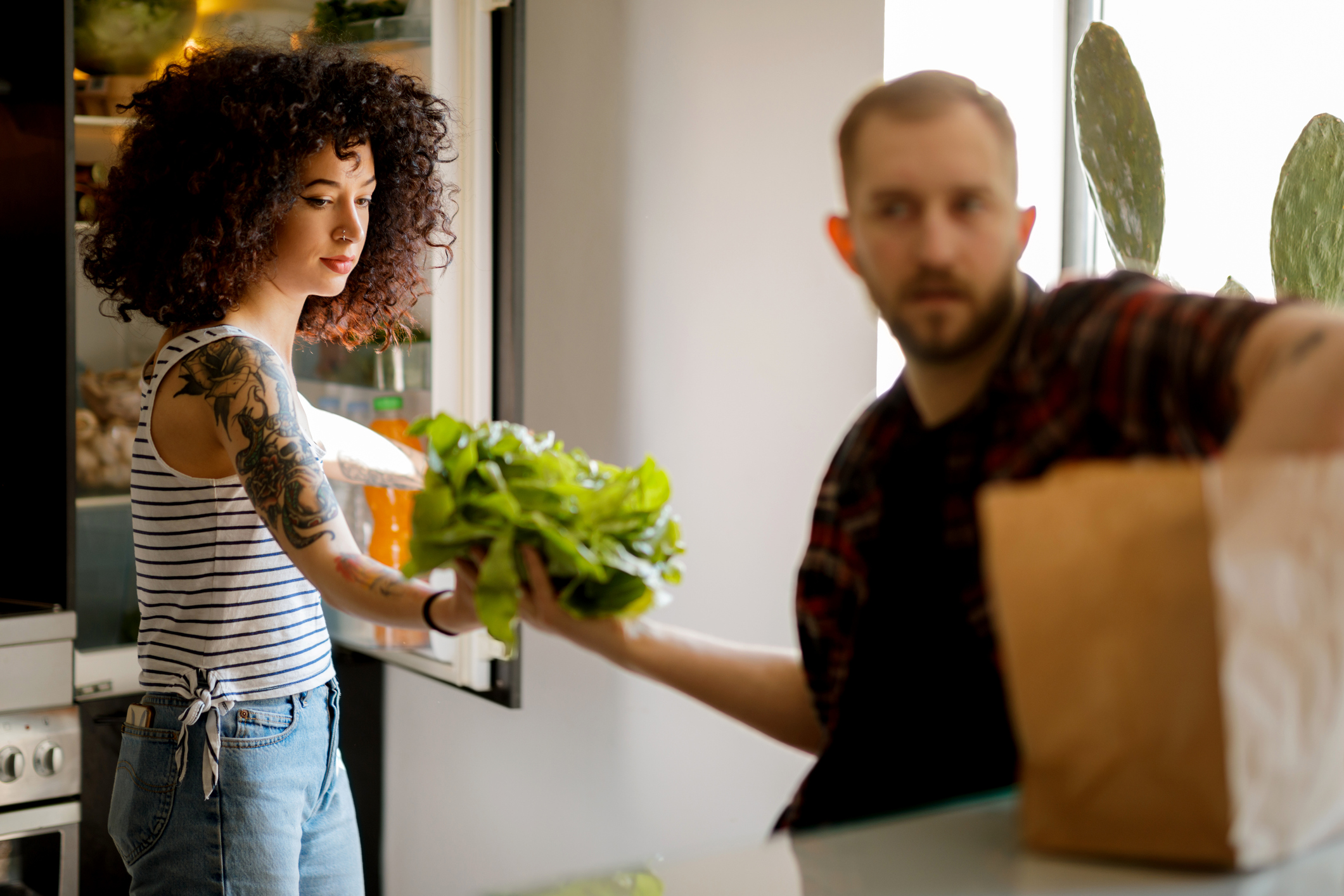Young man passing lettuce and other groceries to female roomate opening the fridge