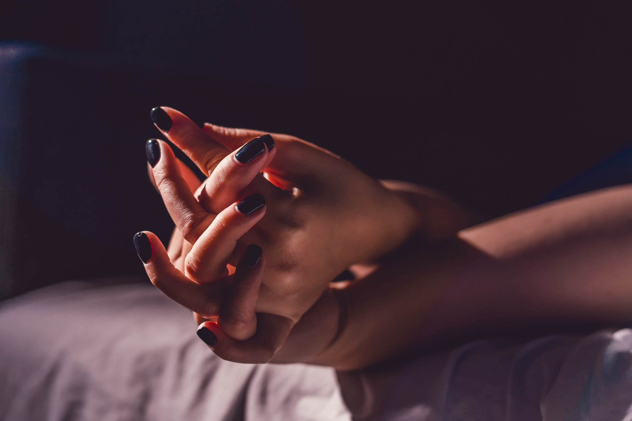 Close up on female young woman's girl's beautiful hands with black nail polish in dark room holding crossed fingers on the bed sheet.