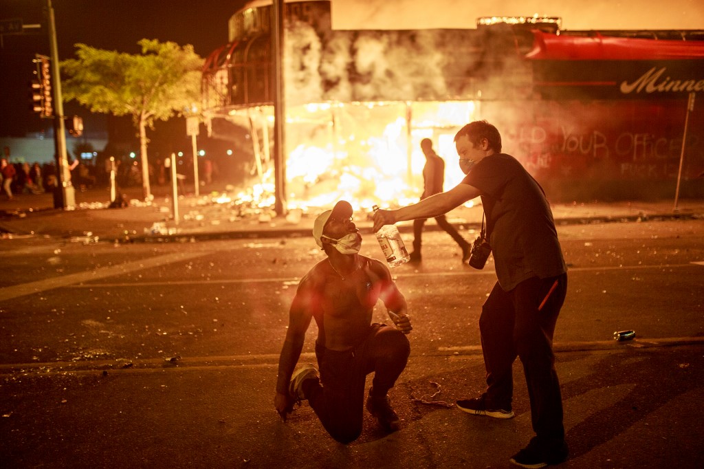 A protester pours vodka into the mouth of another in front of a liquor store in flames on May 28, 2020 in Minneapolis, Minnesota, during a protest over the death of George Floyd, an unarmed black man, who died after a police officer kneeled on his neck for several minutes. - A police precinct in Minnesota went up in flames late on May 28 in a third day of demonstrations as the so-called Twin Cities of Minneapolis and St. Paul seethed over the shocking police killing of a handcuffed black man. The precinct, which police had abandoned, burned after a group of protesters pushed through barriers around the building, breaking windows and chanting slogans. A much larger crowd demonstrated as the building went up in flames. (Photo by Kerem Yucel / AFP)