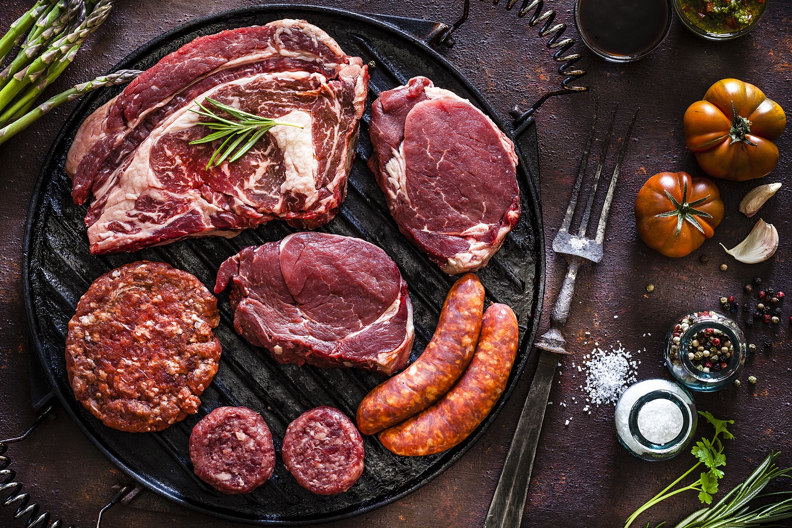 Various cuts of raw meat shot from above on rustic kitchen table. The cuts are on a cast iron grill and includes Angus steak, tenderloin, sausages and hamburger meat. The grill is surrounded by herbs, vegetables and spices for cooking meat like peppercorns, salt, garlic, tomatoes, asparagus and rosemary. A vintage fork is included in the composition. Predominant colors are red and brown. Low key DSRL studio photo taken with Canon EOS 5D Mk II and Canon EF 100mm f/2.8L Macro IS USM.