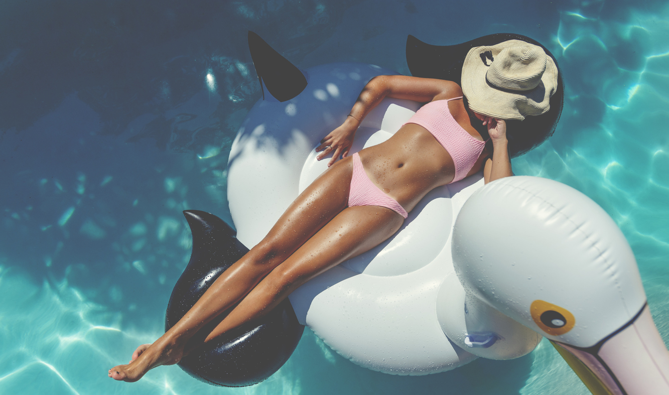 Woman floating on a white inflatable in swimming pool in a pink bikini. She is tanned in turquoise water wearing a sun hat.