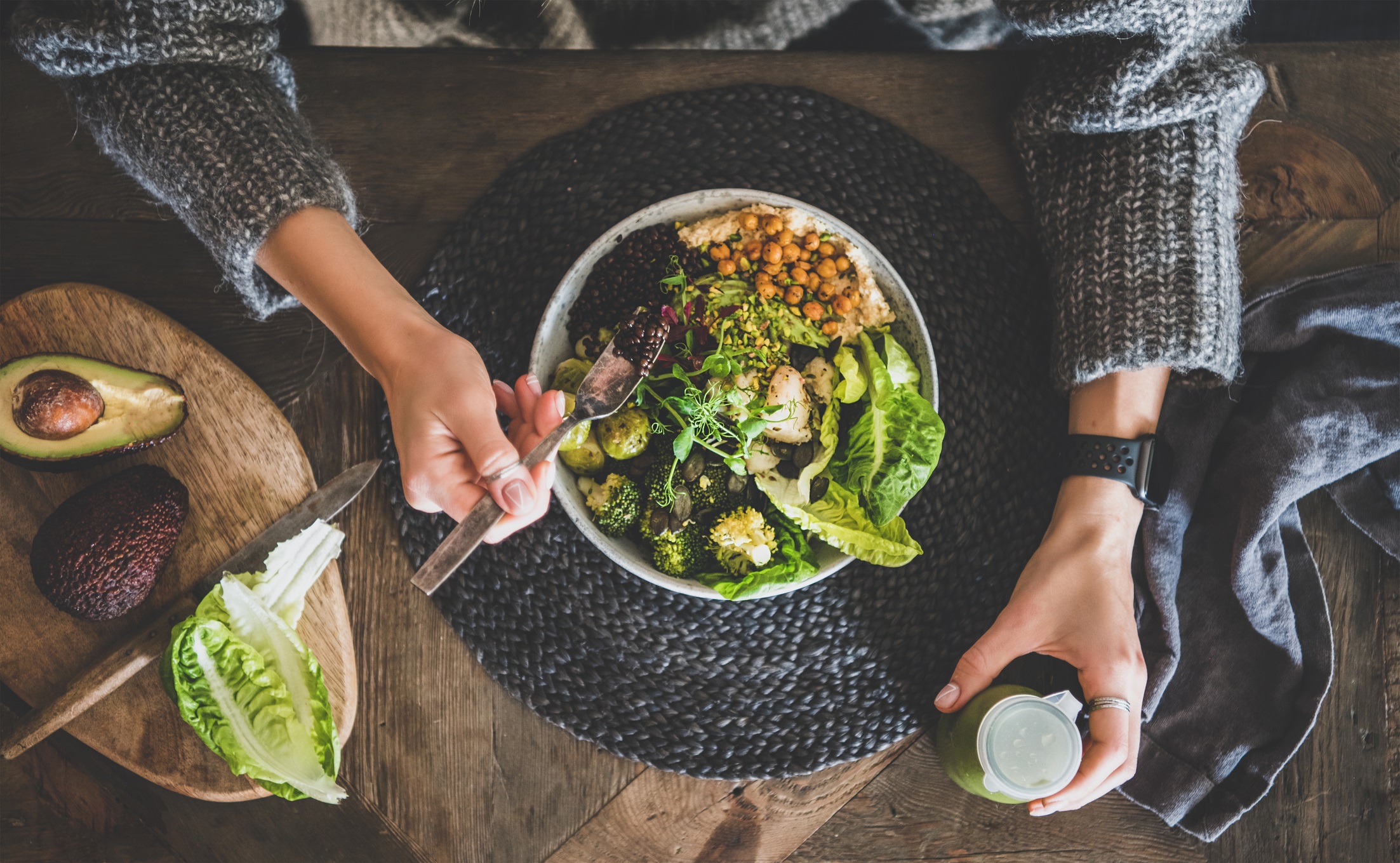 Healthy dinner, lunch setting. Flat-lay of vegan superbowl or Buddha bowl with hummus, vegetable, salad, beans, couscous and avocado, green smoothie and woman's hands over wooden table, top view