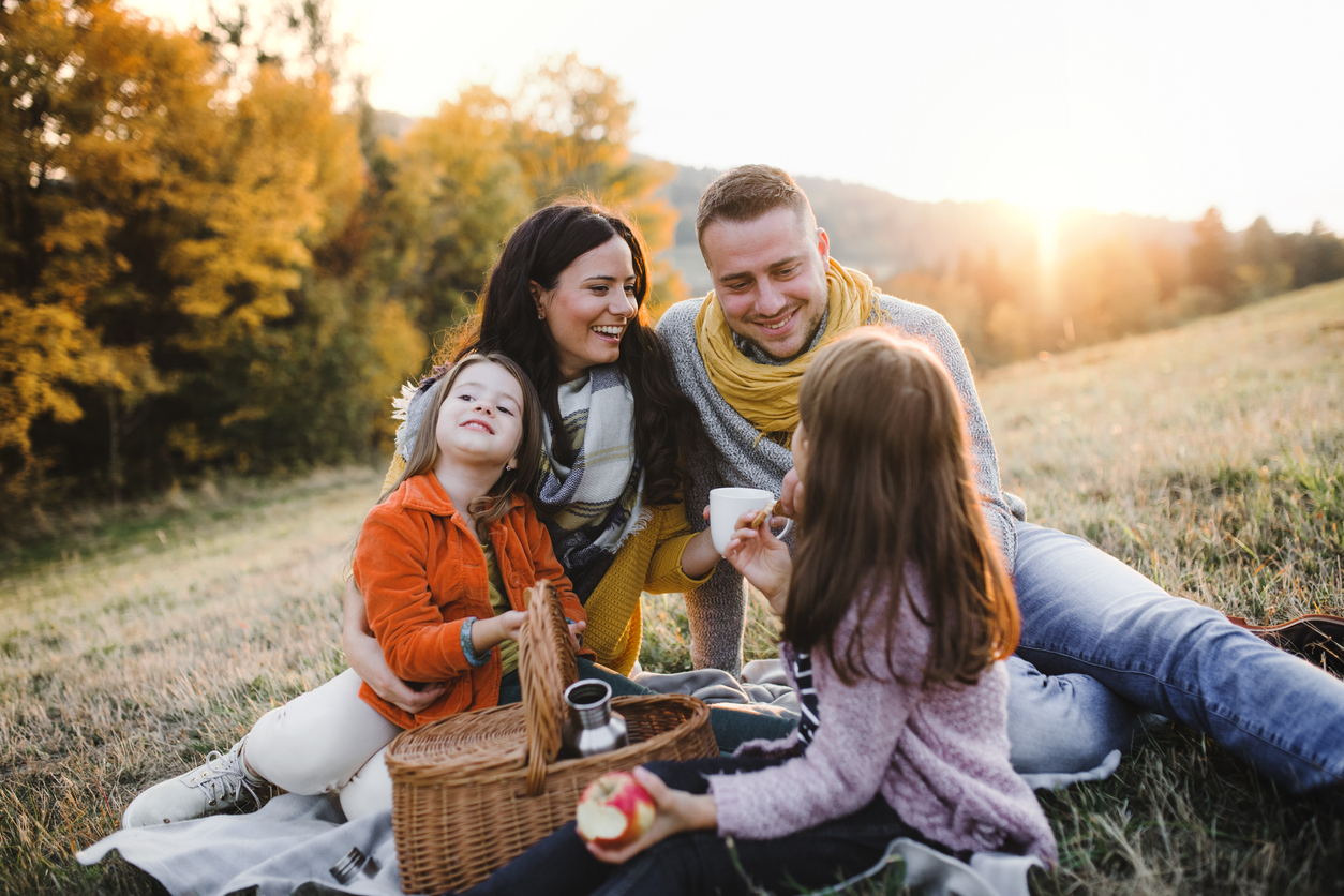 A portrait of happy young family with two small children sitting on a ground in autumn nature at sunset, having picnic.