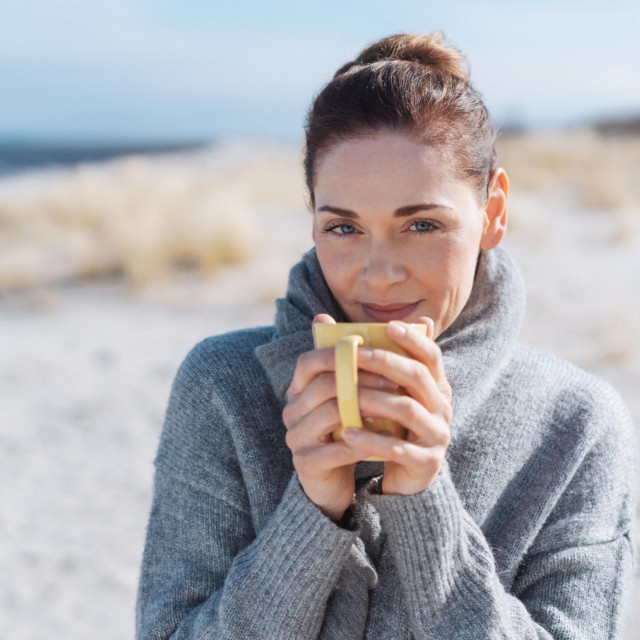 Smiling woman relaxing with coffee at the seaside standing on a sandy beach in a warm winter scarf smiling at the camera
