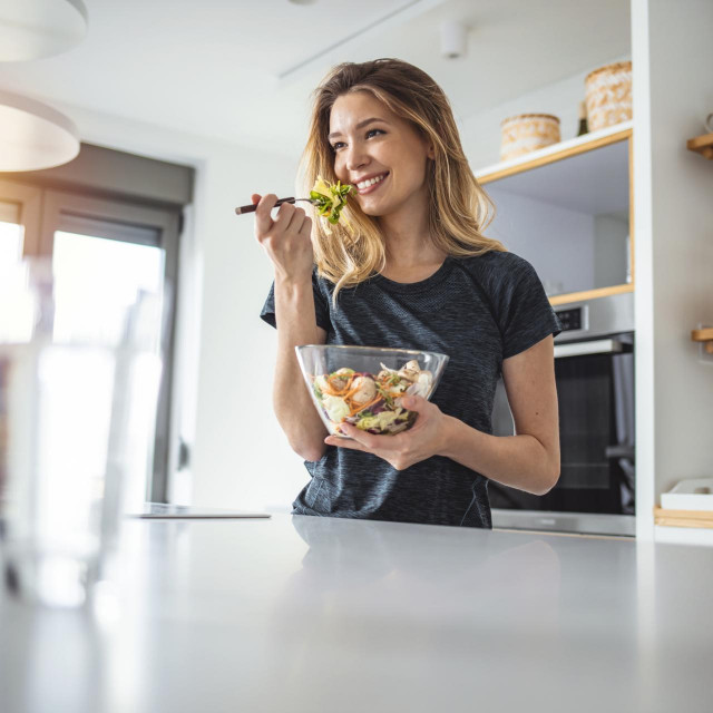 Photo of young woman enjoying a delicious salad while standing in her kitchen at home during the day.