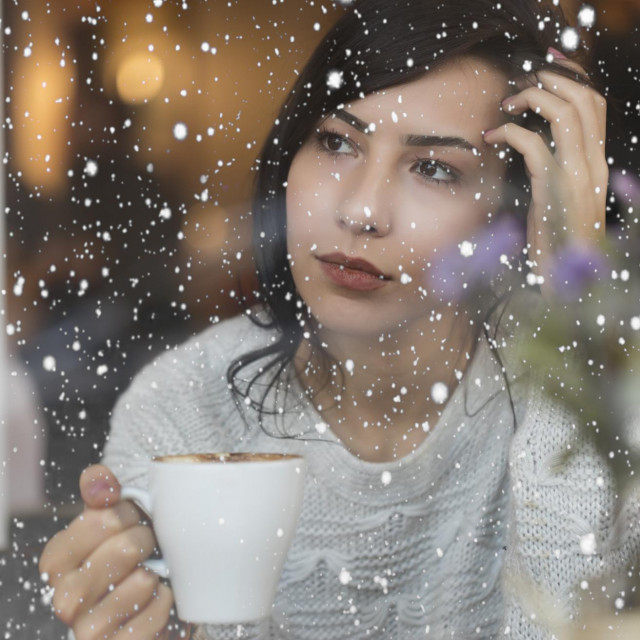 Beautiful Young Woman drinking coffee and watching snow through the window