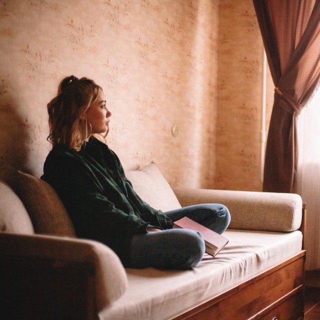 Young sad woman holding book looking through window while sitting on sofa in living room at home