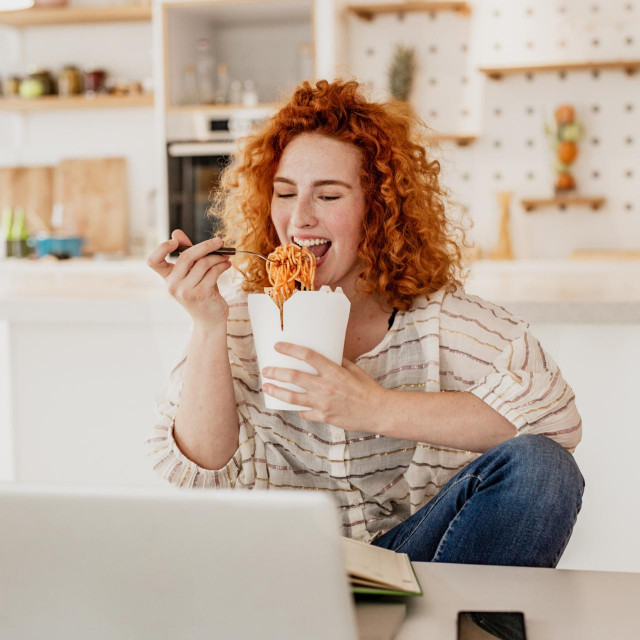 A young woman is using a laptop at home, she is sitting at the table and eating takeaway pasta and watching something on the laptop
