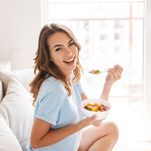 Cheerful young woman eating healthy breakfast while sitting on a couch at home