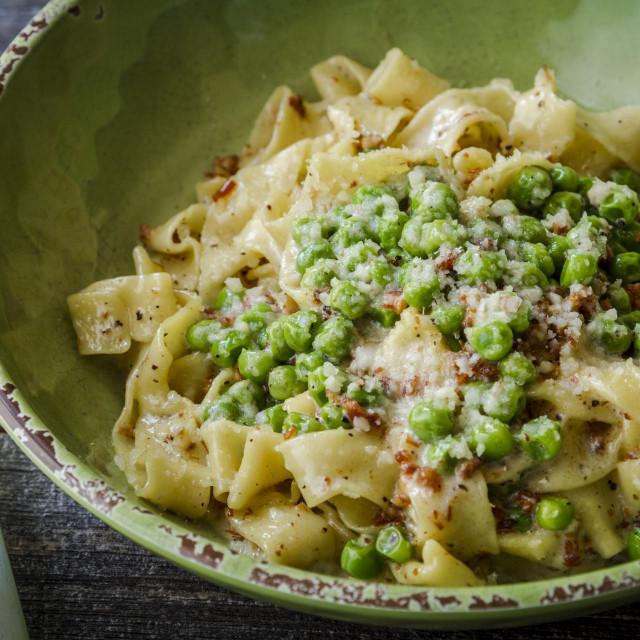 Fresh Carbonara with fresh green peas, pancetta, creamy sauce with fettuccine noodles garnished with fontina cheese in rustic green bowl with fork on side
