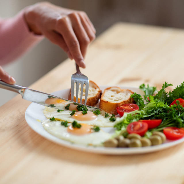 Woman eating tasty breakfast in kitchen, having plate with eggs, bread, olives and salad. Unrecognizable female using fork and knife while enjoying her delicious meal, cropped image, closeup