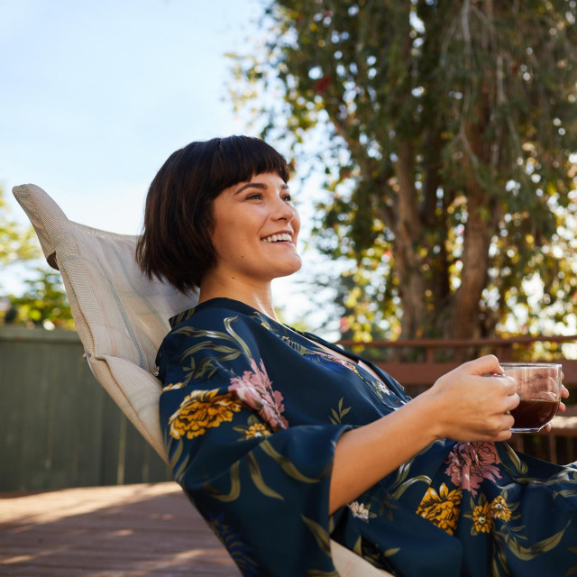 Smiling young woman wearing a robe and lying on a deck chair outside drinking a cup of tea