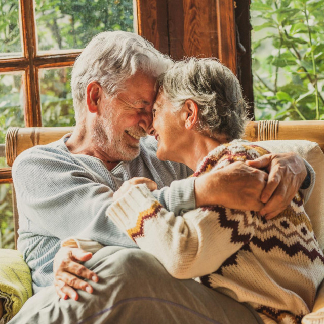 Old people senior man and woman in love and tenderness at home. Mature elderly couple enjoy relationship hugging and caring each other sitting on the sofa in living room with outdoor view
