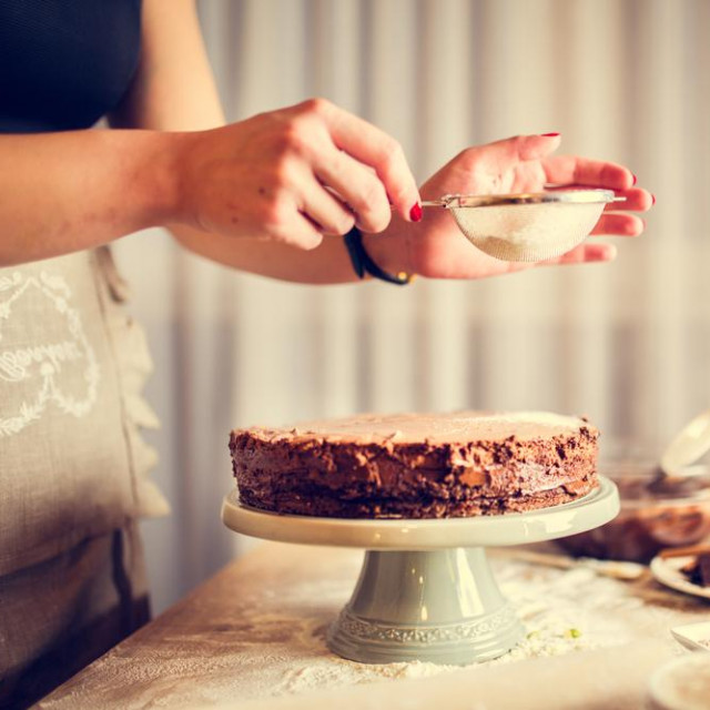 &lt;p&gt;House wife wearing apron making finishing touches on birthday dessert chocolate cake.Woman making homemade cake with easy recipe,sprinkling powdered sugar on top.Icing sugar sprinkled with colander&lt;/p&gt;

