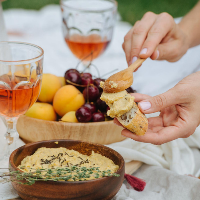&lt;p&gt;Woman hand smearing hummus on bread with a spoon. It‘s taken from the wooden bowl with thyme branch lying on its edge. Next to it is wine glass and bowl with cherries, apricots and pizza.&lt;/p&gt;