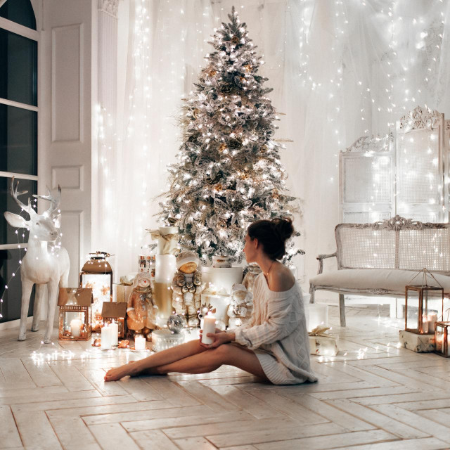 &lt;p&gt;woman in white sweater, warm and cozy evening in Christmas interior design, Xmas tree decorated by lights gifts toys, candles, lanterns, garland lighting indoors fireplace.holiday living room.New year&lt;/p&gt;