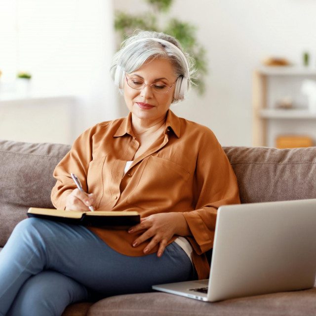 &lt;p&gt;Elderly female in headphones and glasses writing in notebook and listening to music while sitting on couch near laptop&lt;/p&gt;