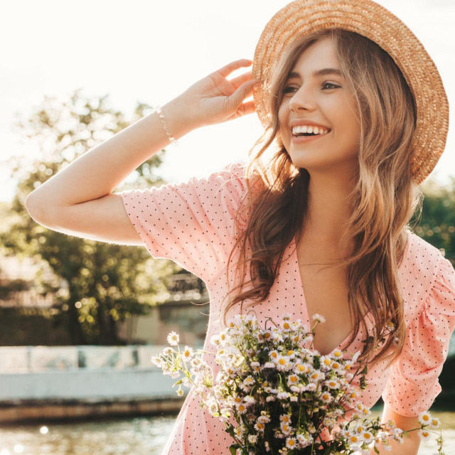 &lt;p&gt;Young beautiful smiling hipster woman in trendy summer sundress. Sexy carefree woman posing on the street background in hat at sunset. Positive model outdoors on embankment. Holding flowers in hands&lt;/p&gt;