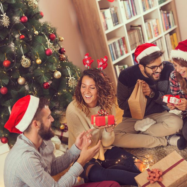 Group of young friends sitting next to a nicely decorated Christmas tree, exchanging Christmas presents. Focus on the girl wearing antlers