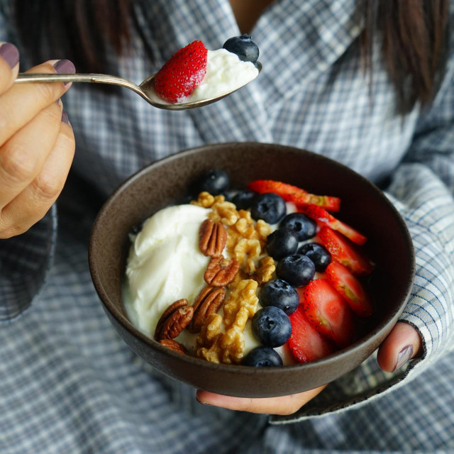 &lt;p&gt;Probiotic and ketogenic food theme bowl of greek yogurt topped with pecan, walnut, blueberries, strawberries in female left hand and right hand holding silver spoon with yogurt and fresh berries.&lt;/p&gt;