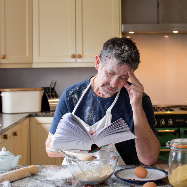 Man looks at a cookery book in the kitchen. Wearing an apron, surrounded by ingredients and mess. Learning how to bake is a new challenge for a mature man. Real life skills concept. Cookery school.