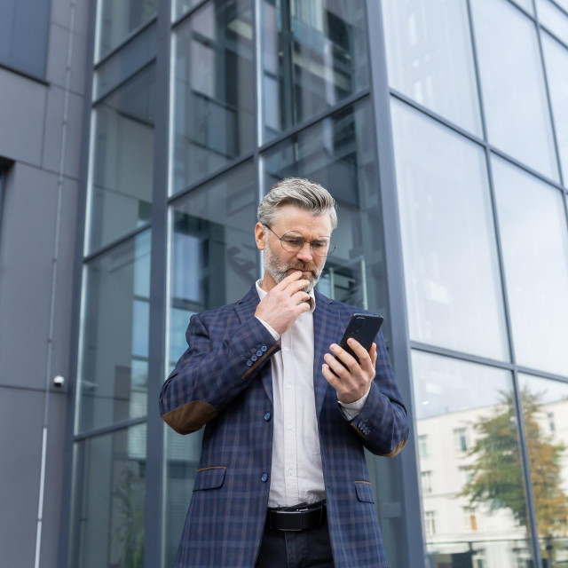 An older gray-haired man in a suit and glasses stands outside an office, a skyscraper. Worriedly looks at the phone, received a message, reads, calls.
