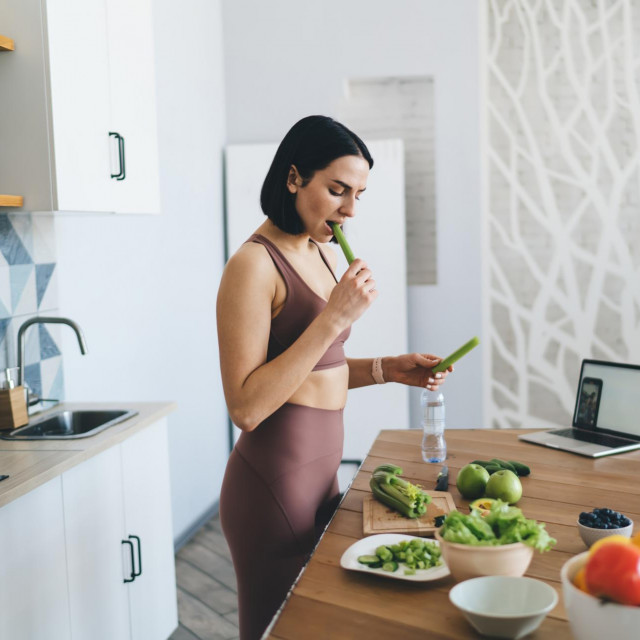 Side view of focused young female in sportswear with mobile and netbook biting celery while cooking healthy salad at wooden table in contemporary kitchen