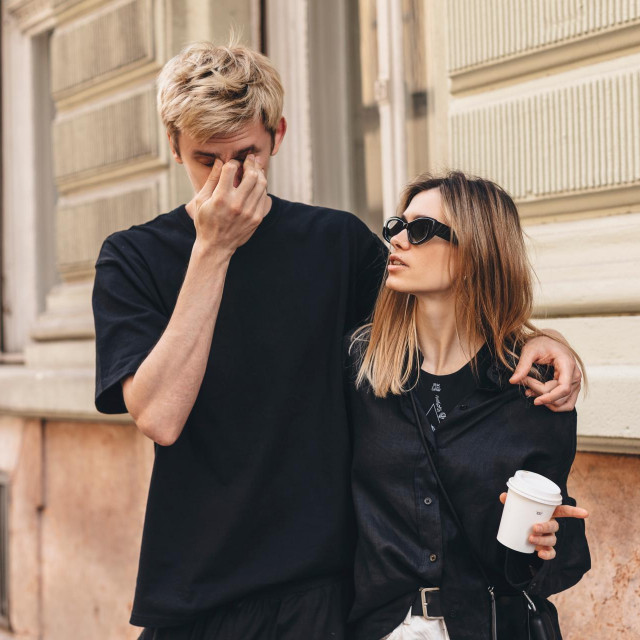 Stylish couple in love hugging walk on the street in old city. Modern woman hold coffee cup and look at man in fashionable black clothes. Man feel bad, holding bridge of nose and suffering in pain.