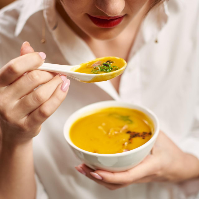 Crop of woman with red lips in white blouse holding white bowl of orange cream soup. Client tasting delicious and delicate meal in luxury restaurant. Girl with manicure eating first dish on dinner.