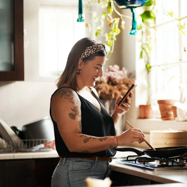 Happy, woman cooking food with smartphone and in kitchen of a home. Dinner or lunch, multitasking and plus size, natural person with tattoo on cellphone following recipe motivation for healthy diet