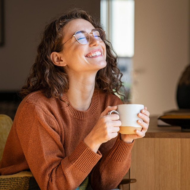 Portrait of joyful young woman enjoying a cup of coffee at home. Smiling pretty girl drinking hot tea in winter. Excited woman wearing spectacles and sweater and laughing in an autumn day.