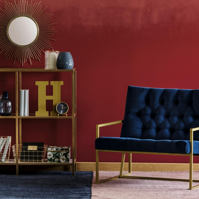 Golden metal rack with books and decor standing in burgundy room interior with navy blue armchair and potted plant