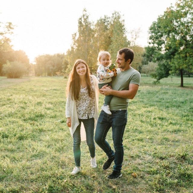 Child embrace parents happiness playing in green grass in park. Happy mother, father hug baby son walking in garden at sunset. Family spending time together outdoors. Children‘s day. Friendly family.