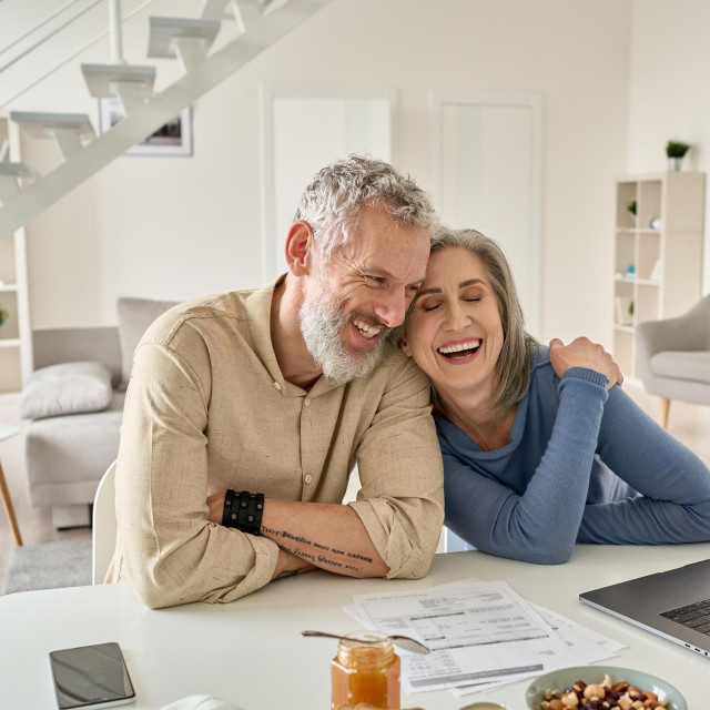 Happy mature older family couple laughing, bonding sitting at home table with laptop. Smiling middle aged senior 50s husband and wife having fun satisfied with buying insurance, paying bills online.