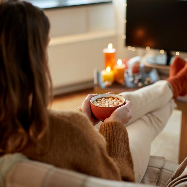 halloween, holidays and leisure concept - young woman watching tv and drinking hot chocolate with marshmallow with her feet on table at cozy home