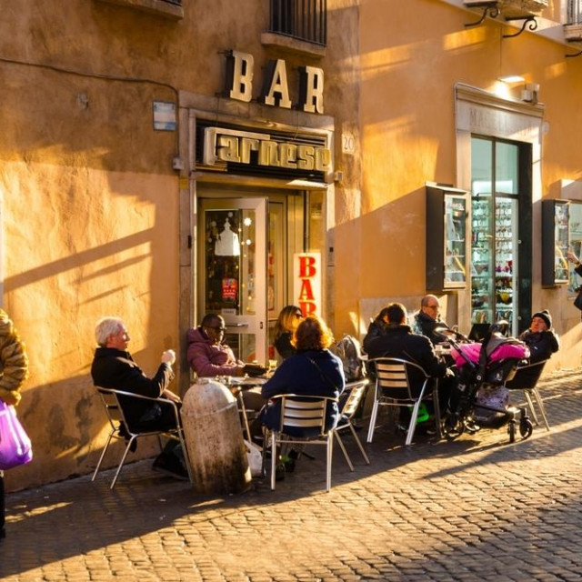 Rome, Italy, January 3rd, 2020Italian people having antipasti and cafe sitting around small table on chairs in front of a caffee bar in a warm winter evening light casting long shadows