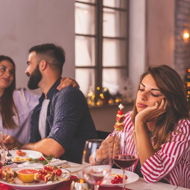 Lonely woman spending Christmas Eve with friends at home, sad while sitting over Christmas dinner surrounded with couples in love