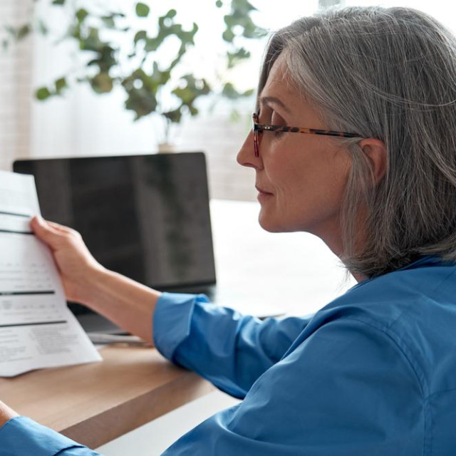 Senior mature business woman holding paper bill using calculator, old lady managing account finances, calculating money budget tax, planning banking loan debt pension payment sit at home office table.