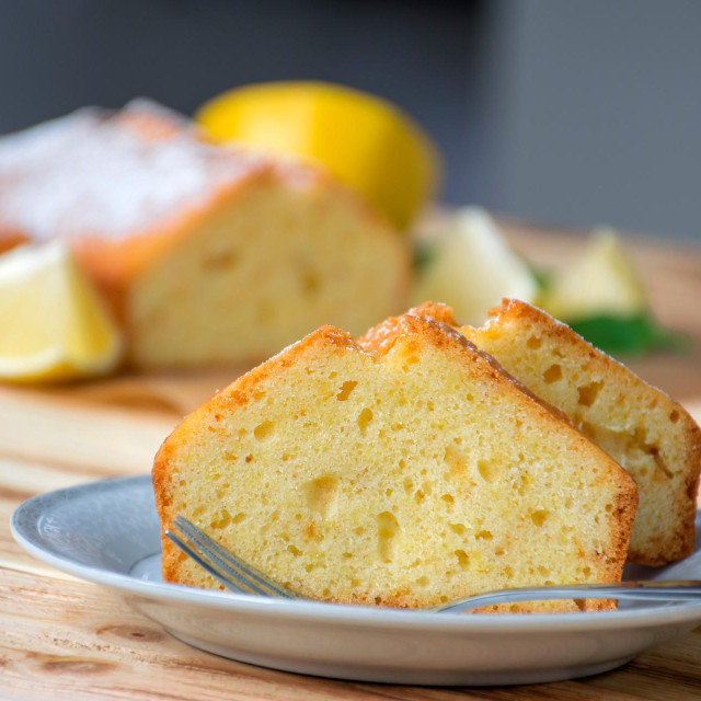 Piece of lemon cake on plate on rustic wooden board with full pie and lemons on background. Easy recipe of citrus dessert for everyday cooking. Homemade bakery by classic recipe.