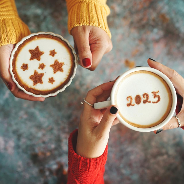 New year 2025 celebrated coffee cup with the number 2025 on frothy surface in female hands holding over rustic blue background and another one with stars symbol on frothy surface. Holidays food art.