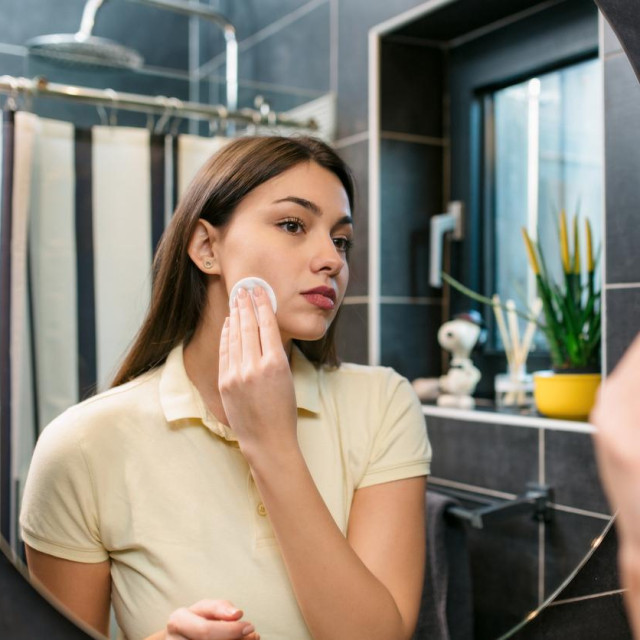 Young Caucasian woman in her bathroom, looking herself in a mirror and taking makeup off her face